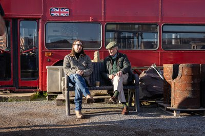 "Daniel Dawson Gordon and Jeremy Hackett sit on a slate bench in front of a red London Double Decker Bus"